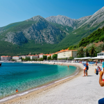 **

"A scenic view of the Montenegrin coast, featuring a modern resort with fully clothed tourists enjoying the beach, surrounded by mountains. In the background, a glimpse of a port with cargo ships. Safe for work, appropriate content, family-friendly, professional photography, high quality, perfect anatomy, natural proportions, well-formed hands, proper finger count, natural body proportions."

**